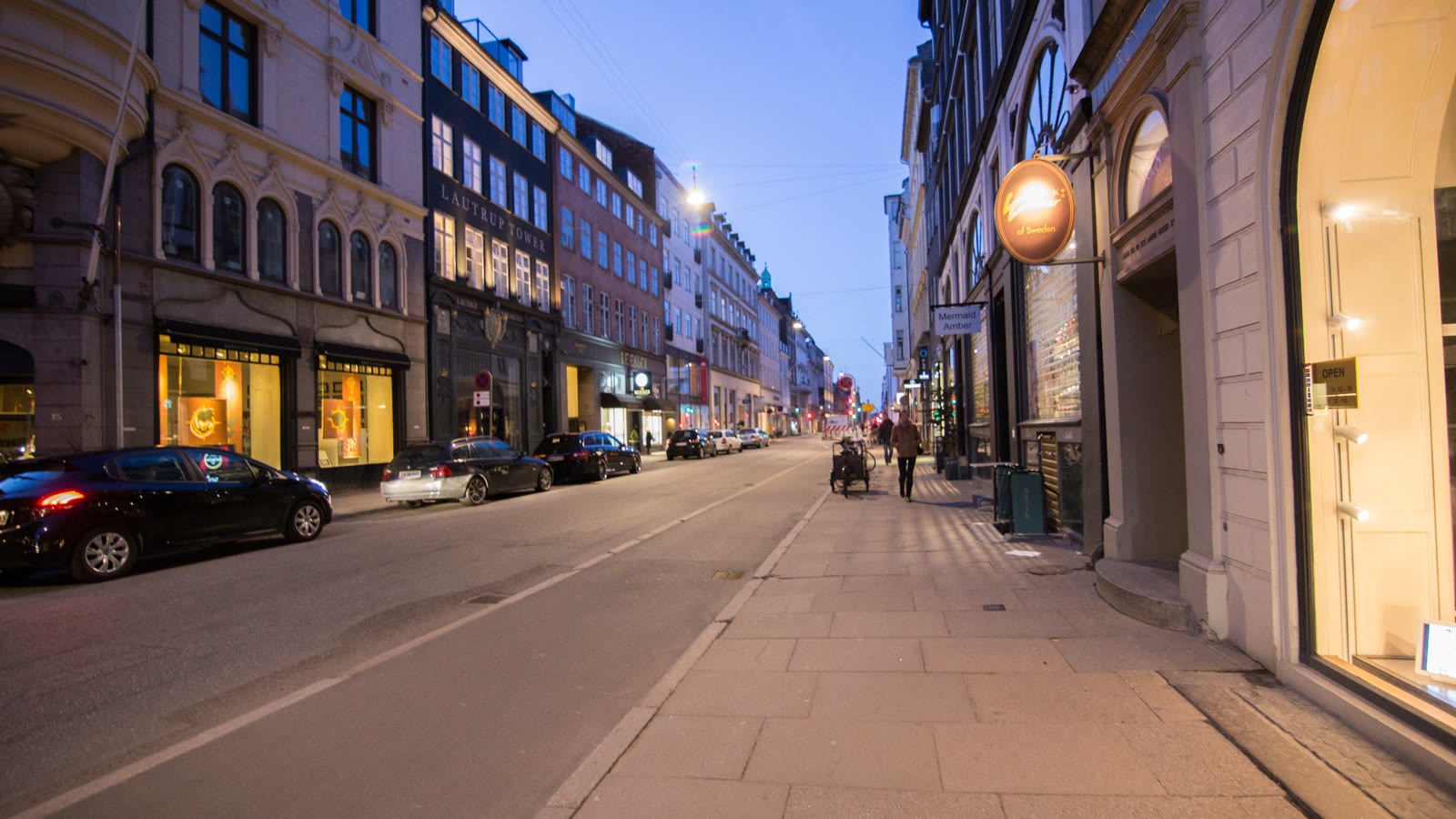 Bredgade i skumringen med oplyste butiksvinduer paa begge sider af gaden